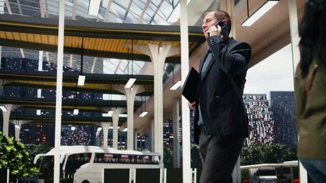 Businessman makes a phone call while walking through a bus terminal before a work trip; the business traveler manages mobile communication in the coach station transit area before departure.