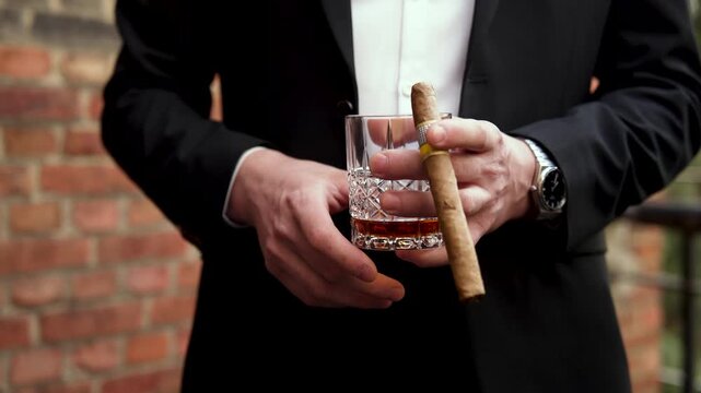 Businessman in a luxury suit holding a crystal glass of whiskey and a premium cuban cigar close-up.