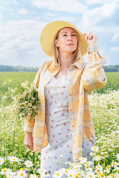 Blonde woman wearing straw hat, floral dress, and yellow plaid shirt stands in field of daisies, holding bouquet of wildflowers in her hand, cradling her hat and gazing into distance