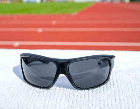 Black sports sunglasses sitting on a white textured towel with a blurred running track behind.