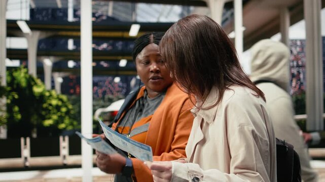 Travel companions read boarding passes together in a bus station while discussing the journey; the women compare bus ticket information in the shuttle terminal.