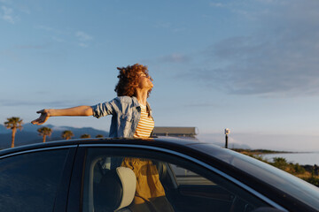 Fototapeta premium A woman with curly hair stands through an open car window along a sunny coastline, arms outstretched. A lifestyle moment of freedom, travel, and optimistic daydreams at the seaside.