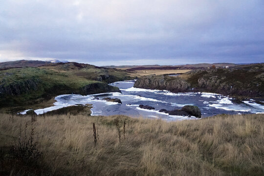 An icy river winds through a rugged landscape of grassy hills and rocky bluffs under a cloudy twilight sky near Borgarnes, Iceland
