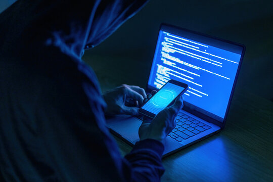 A hooded individual uses a smartphone and a laptop displaying code in a dark, tech-focused setting.