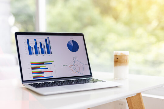A laptop showing data charts sits on a desk next to an iced coffee, with outdoor greenery visible in the background.