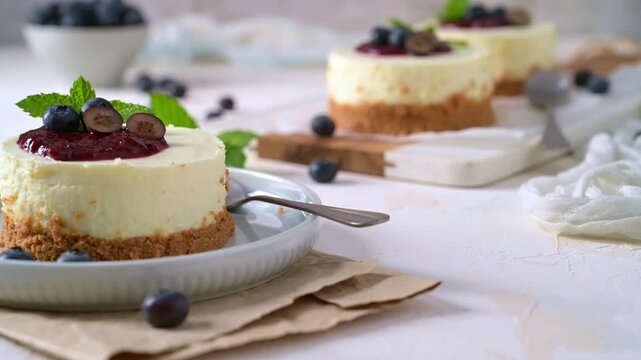 Mini cheesecakes topped with vibrant berry compote and fresh mint are illuminated by soft natural light on a rustic white background with scattered blueberries