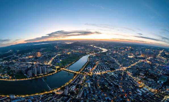 A high-angle aerial panorama of a city with a winding river, captured at dusk with illuminated buildings and roads.