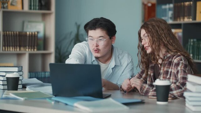 Two students collaborate on a laptop at a desk with books and notebooks