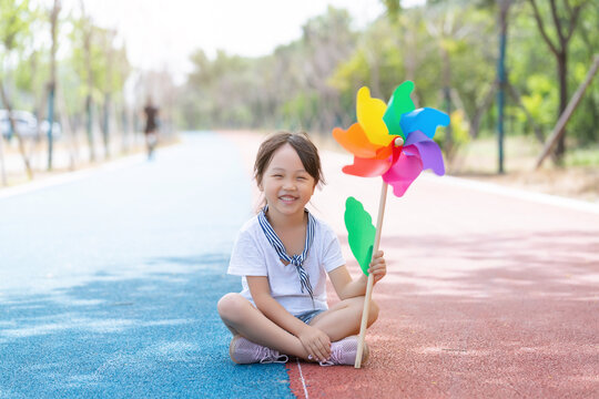 Cute little girl sitting cross-legged on a colorful pathway with a large pinwheel in hand.