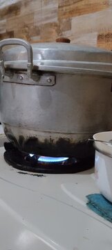 A close-up shot of a large, used aluminum stock pot with a lid, sitting on a lit gas stove burner in a home kitchen. The bottom of the pot shows signs of soot and wear from regular cooking, capturing 