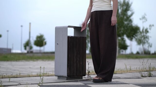Woman in casual outfit throws empty plastic water bottle into outdoor metal and wood trash bin on paved path with trees and weeds under cloudy sky.