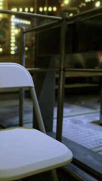 A white folding chair sits quietly in a lively carnival atmosphere, surrounded by sparkling lights and a festive ambiance. Joyful sounds echo as people enjoy the evening.