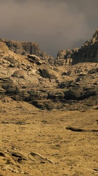 Barren desert expanse stretches into the distance with rugged rocks rising sharply. Dark clouds loom above, casting shadows on the arid ground and enhancing the stark beauty of the scene.