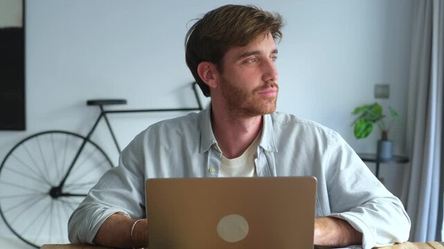 Thoughtful young man working remotely on a laptop computer. Then looking away and smiling confidently at the camera while sitting at his desk in a modern home office with a bicycle in the background
