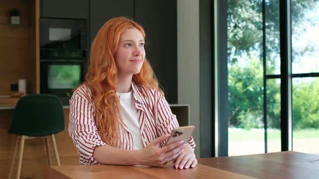 Thoughtful young woman with long red hair sitting at a table, using her smartphone and looking concerned before her expression changes to a happy, pensive smile while looking away