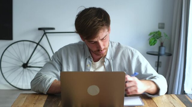 Young handsome man attentively watching a webinar on his computer and taking notes in a notebook. Millennial student learning remotely from home, engaged in e-learning and online education