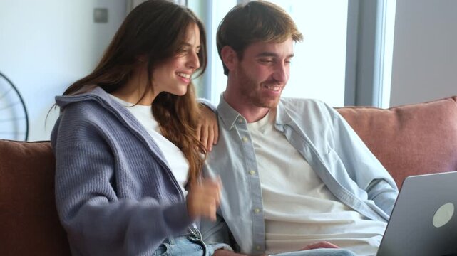 Cheerful young couple sitting together on the couch, using a laptop for a video call with friends or family, smiling and waving at the screen in their cozy living room at home