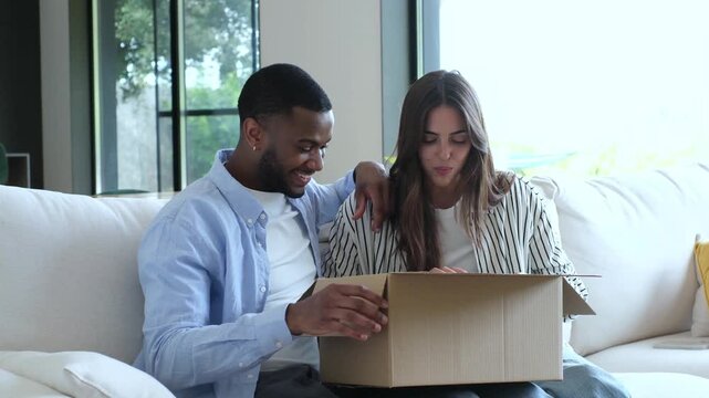 Young, excited, multiethnic couple sitting together on a sofa at home, eagerly opening a cardboard box and expressing surprise and happiness at the contents of their online shopping order