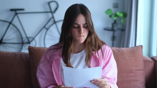 Worried and upset young woman sitting on the couch at home, reading a letter containing bad news, feeling stressed and frustrated by the unexpected information about debt or eviction