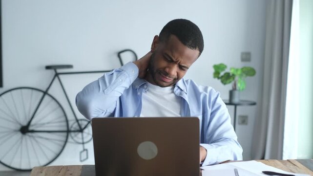 Overworked african american freelancer suffering from neck pain while working on a laptop at his desk, experiencing discomfort from a sedentary job and trying to relieve muscle tension