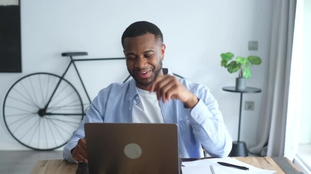 Happy young black male professional using a laptop for a video conference call, smiling and gesturing while working remotely from his modern home office with a bicycle in the background