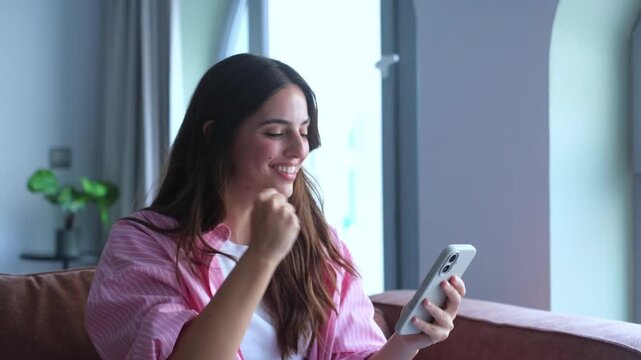 Overjoyed woman sitting on a couch receiving amazing news on her smartphone, celebrating a big win, success, or achievement with a yes gesture, feeling ecstatic and triumphant at home