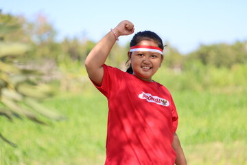 Naklejka premium A girl raises a clenched fist, standing against a backdrop of green trees, wearing a red and white flag ribbon, celebrating Indonesian Independence Day.