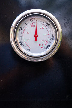 Close up photo of a round metal grill thermometer mounted on a black barbecue lid, showing cooking temperature markings with copy space.