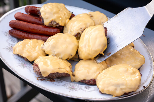 Fresh grilled cheeseburgers and hot dogs on a serving plate with a metal spatula lifting one burger at an outdoor cookout.