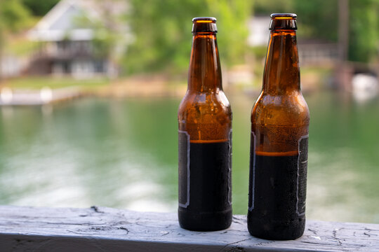 Two condensation-covered brown glass bottles sitting on a wooden railing with a blurred lake and trees in the background.