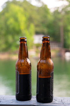 Vertical photo of two chilled brown glass bottles on a wooden railing overlooking a soft blurred lake and green outdoor background.
