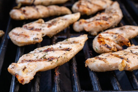 Close-up photo of grilled chicken strips cooking on a barbecue grill with herbs, seasoning, and visible grill marks.
