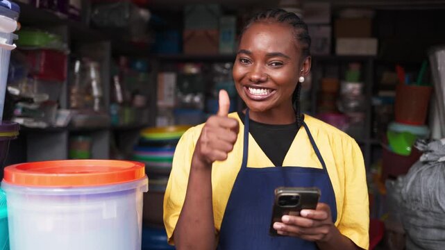 African woman small business owner smiling, standing in a shop, giving a thumbs up while holding smartphone