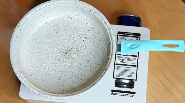 water rapidly boiling in a speckled pan with a blue handle. The saucepan sits on a white portable gas stove with a blue butane canister on an indoor wooden table, emitting white steam.