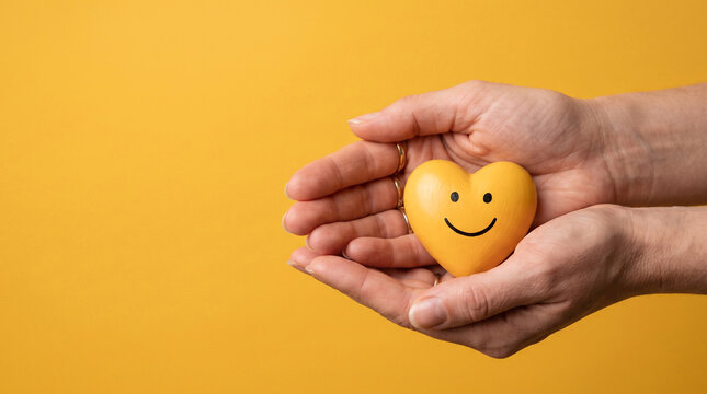 Two hands carefully holding small yellow heart with smiley face on bright yellow background representing happiness mental health and kindness.