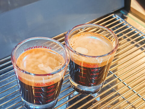 two fresh espresso shots in transparent glass measuring cups placed on a professional coffee machine tray. The image represent coffee preparation, barista culture, cafe beverages, and espresso brewing