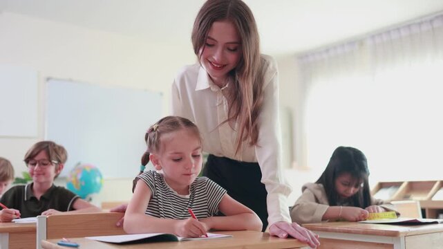 Friendly instructor supporting elementary student during lesson, Teacher helping young student with schoolwork in classroom, Education concept with teacher assisting pupils in classroom