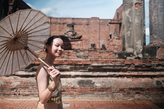 Portrait of Asian teenage girl dressed in traditional Thai outfit and umbrella with cheerful smile at old brick wall, summer holiday travel in Ayutthaya, ancient ruins heritage,  tourist destination.