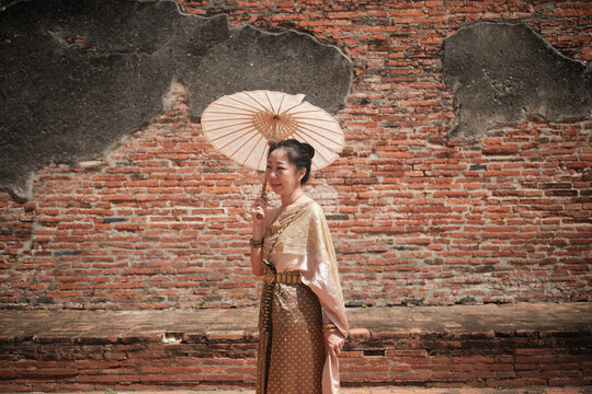 Asian woman dressed in traditional Thai outfit and umbrella with cheerful smiles at old brick wall, summer holiday travel in Ayutthaya, Southeast Asia, ancient ruins heritage,  tourist destination.