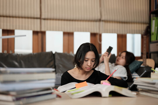 Two Asian teenage tutorials, girl concentrates on learning class at home with pile of homework, her friend uses mobile phone on sofa, self-study with textbooks, homeschooling together lifestyle.