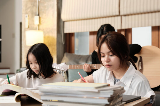 Three Asian teenage tutorials, girls concentrate on reading books for exams, learning at home, her friend relaxes by playing guitar music, self-study with textbooks, homeschooling together lifestyle.