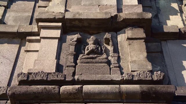 Ancient carved deity sits within a weathered stone niche on the sacred facade of Prambanan Temple.