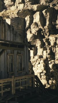 A rustic cabin rests near towering rocky cliffs, bathed in warm golden light as the sun sets. The weathered wooden structure tells stories of past lives among the timeless landscape.