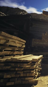 Sunlight illuminates a towering stack of wooden planks at a lumber yard. The warm tones of the wood contrast with the clear blue sky above, creating a serene atmosphere.