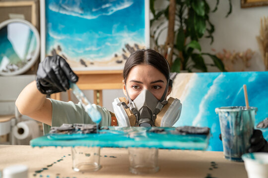 Serious woman artist wearing gloves and respirator working with epoxy resin in creative workshop, female crafter uses spatula tool to add drops of liquid resin on canvas for texture and pattern.