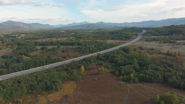 AERIAL: Motorway stretching across the autumn countryside in Croatia. Forest patches, fields, shrubs and distant mountains create a scenic landscape, while light traffic passes along the long roadway