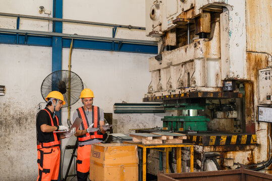 Two engineers in yellow helmets examine a metal strip beside a punch press, one holding a laptop, on the factory floor