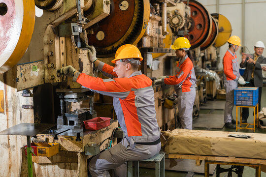 Young factory worker operating mechanical power press machine amid rows of stamping equipment ensuring precision and quality in a modern industrial manufacturing environment with heavy machinery