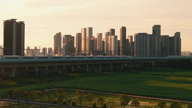 Aerial view of 2 high speed train travel on an elevated track past tall apartment buildings in a city suburb during sunset in China