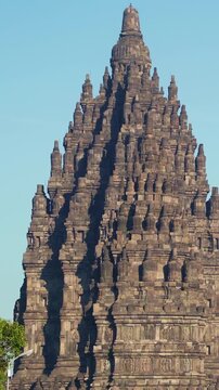 Detailed vertical view of a soaring Prambanan Temple spire reveals intricate stone carvings against a clear blue sky.